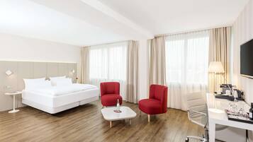 Une chambre d’hôtel avec un lit, deux fauteuils rouges, un bureau avec une lampe et une table basse.