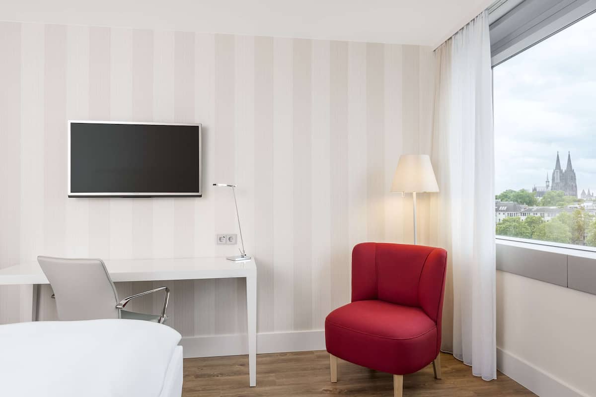 Ein Hotelzimmer mit einem Bett, einem Schreibtisch mit Stuhl, einem Fernseher, einer Lampe und einem Fenster mit Blick auf die Stadt.