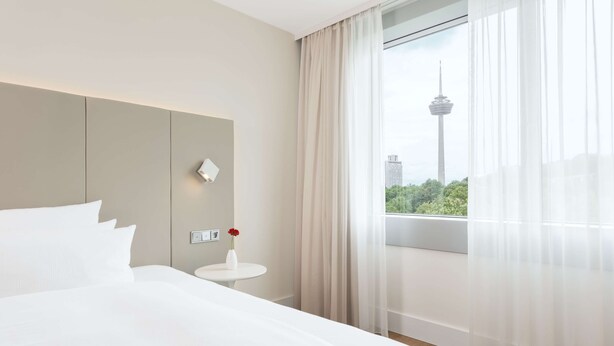 A hotel room with a large bed, a small round table with a vase of flowers, a wall-mounted light, and a window with a view of a tower and greenery. - NH Collection Köln Mediapark (Cologne)