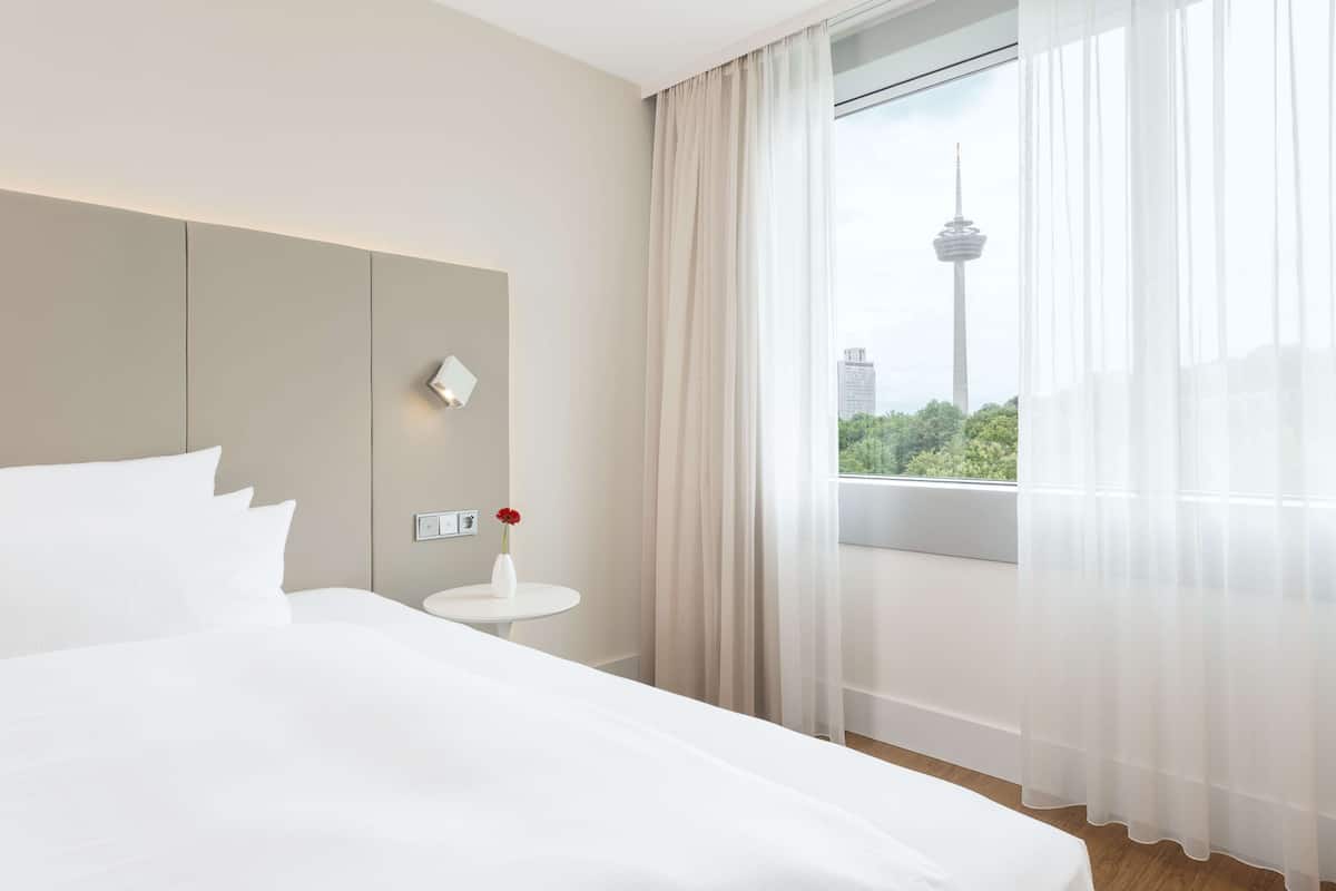 Ein Hotelzimmer mit einem großen Bett, einem kleinen runden Tisch mit Blumen in einer Vase, einer Wandleuchte und einem Fenster mit Blick auf einen Turm und Grünflächen.