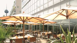 Un espacio al aire libre con mesas y sillas, sombrillas grandes y vistas a un edificio moderno con una torre al fondo.