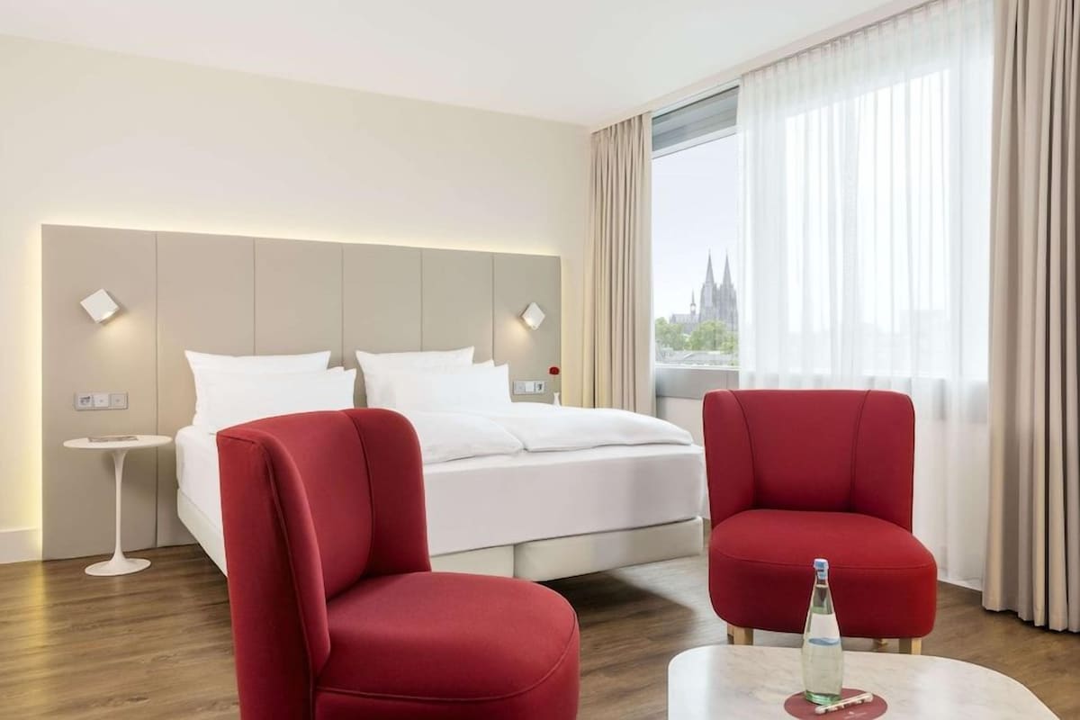 A hotel room with a bed, two red chairs, a small table with a bottle, and a view of a building through sheer curtains.