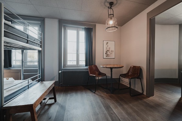 Una habitación con literas, una mesa redonda con sillas, un radiador y una lámpara colgante.