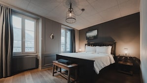 A bedroom with a large bed, a bedside table, a bench, a window, and a radiator. - Falken Hotel & Backpacker (Interlaken)
