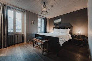 Un dormitorio con una cama grande, una mesita de noche, un banco, una ventana y un radiador.