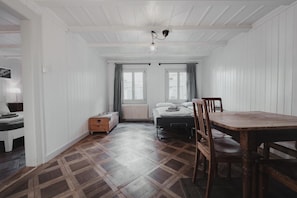 Una habitación con una cama, un cofre de madera, una mesa con sillas y un ventanal con cortinas.