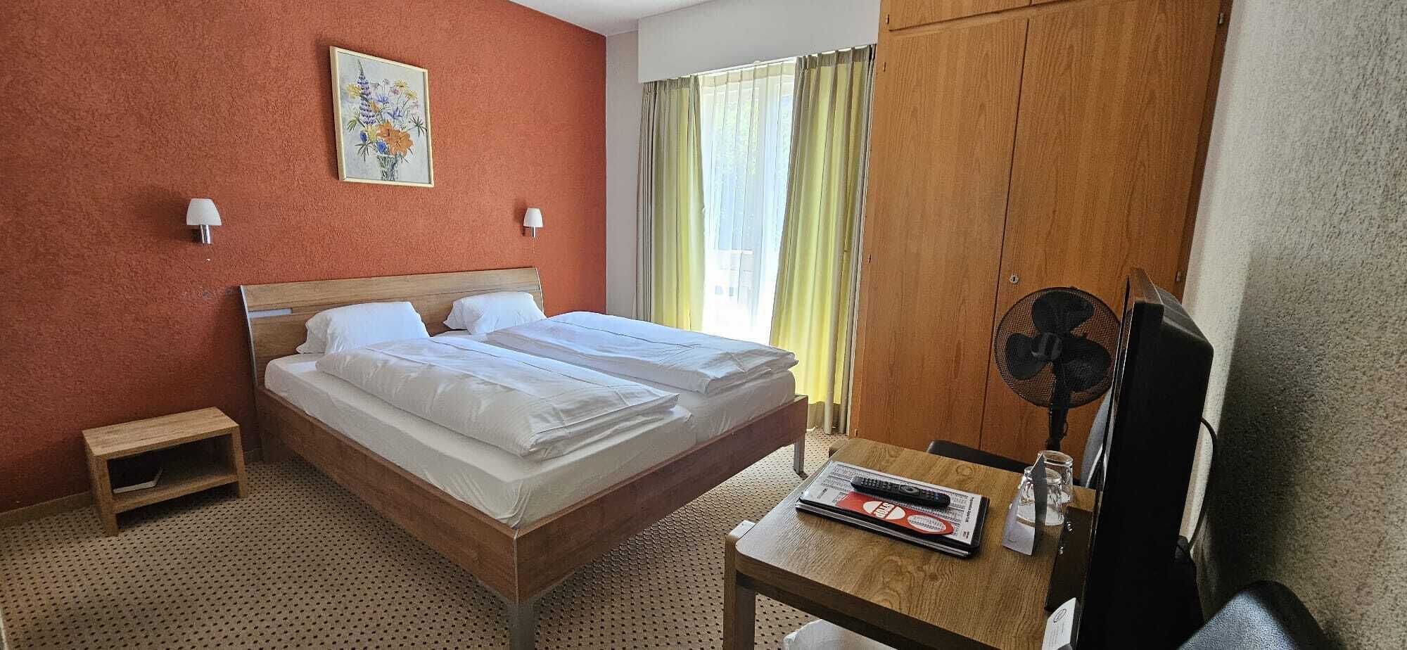 Habitación de hotel con cama doble, una mesita de noche, una silla, escritorio con ventilador y ventana con cortinas.