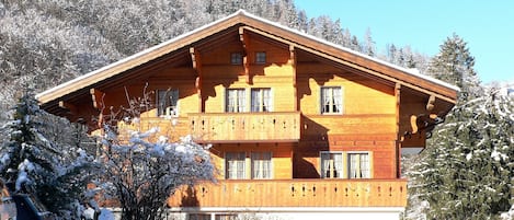 A wooden chalet with a balcony, surrounded by snow-covered trees and a clear sky.