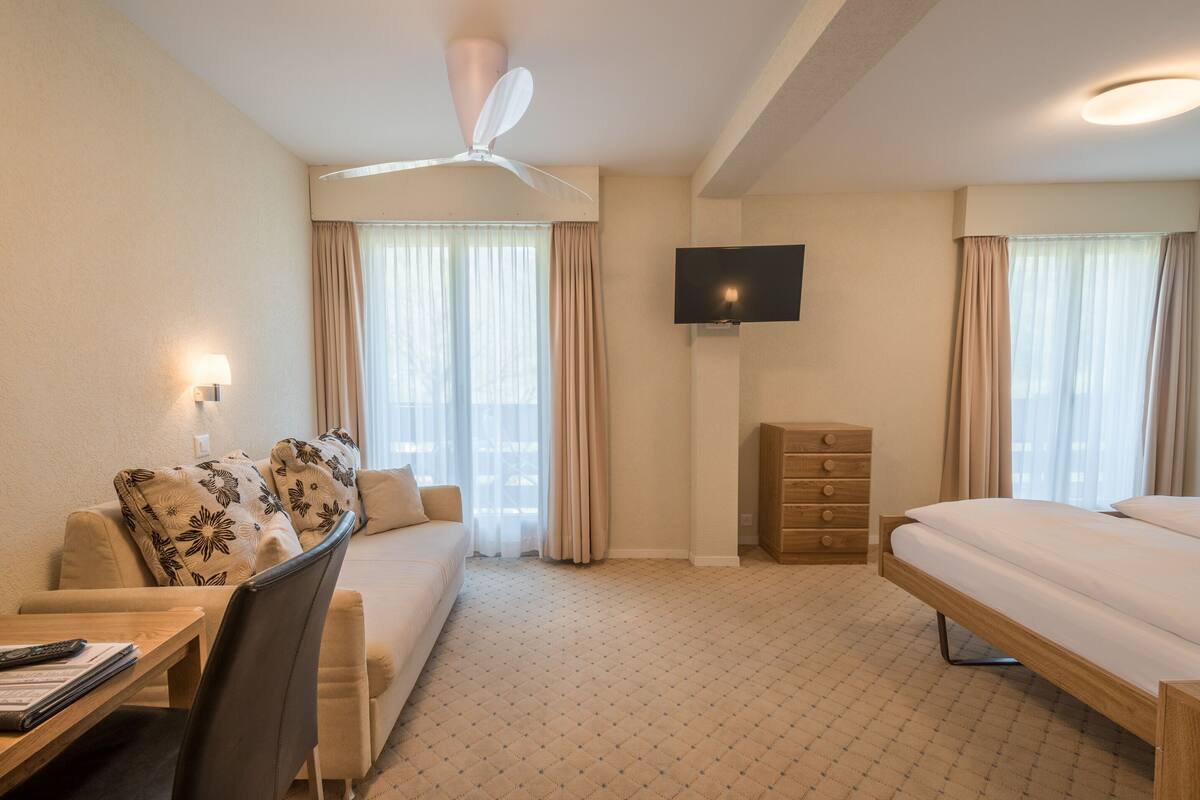 Habitació d'hotel amb un llit, un sofà, un escriptori i una cadira, un televisor fixat a la paret i un ventilador de sostre.
