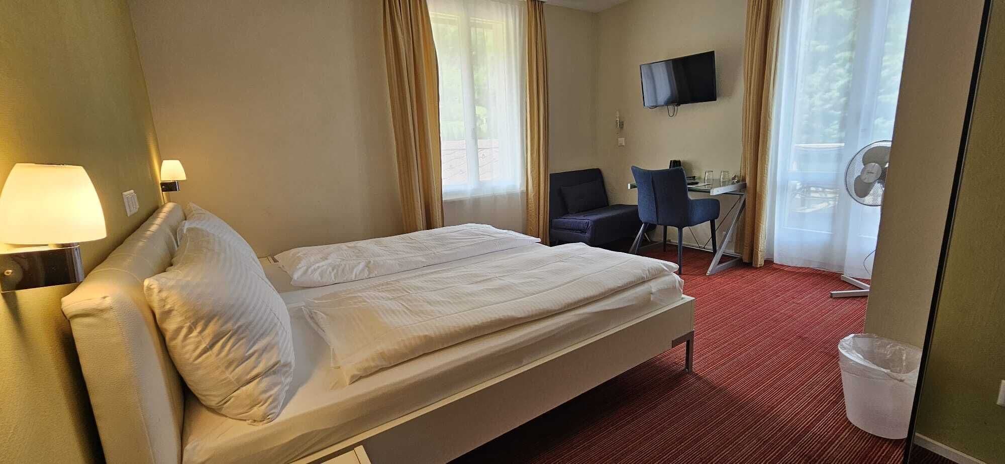 Habitación de hotel con cama, escritorio, silla, televisor y ventana con cortinas.