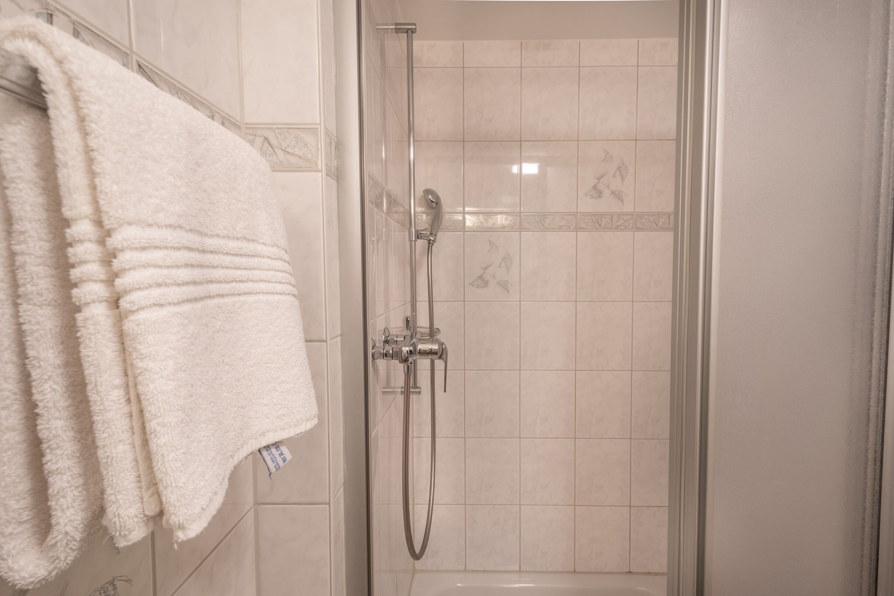 Eine Dusche mit Glastür, ein Handtuchhalter mit gefalteten Handtüchern und Fliesenwände.