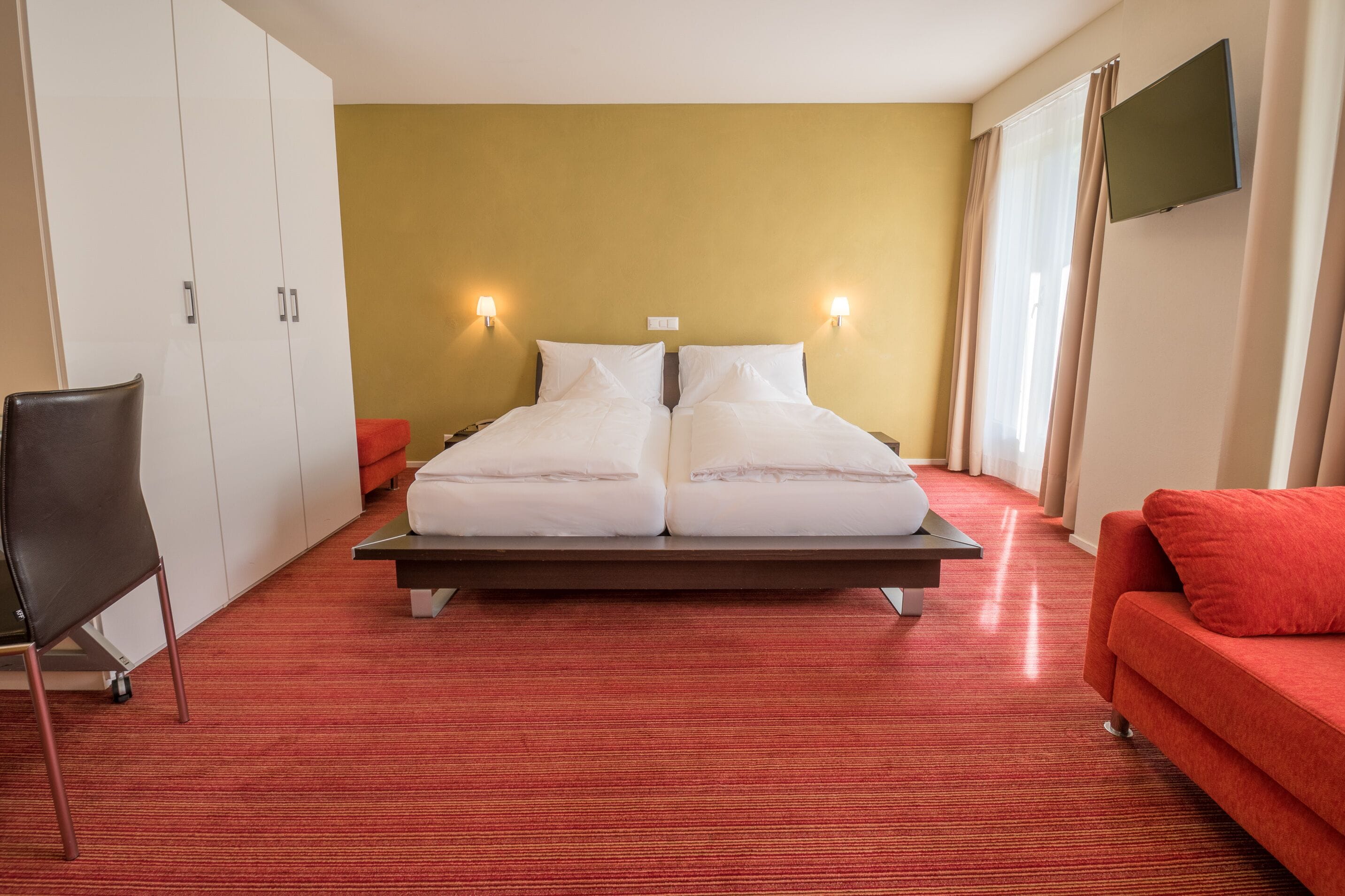A hotel room with a double bed, a red sofa, a TV, and a wardrobe.