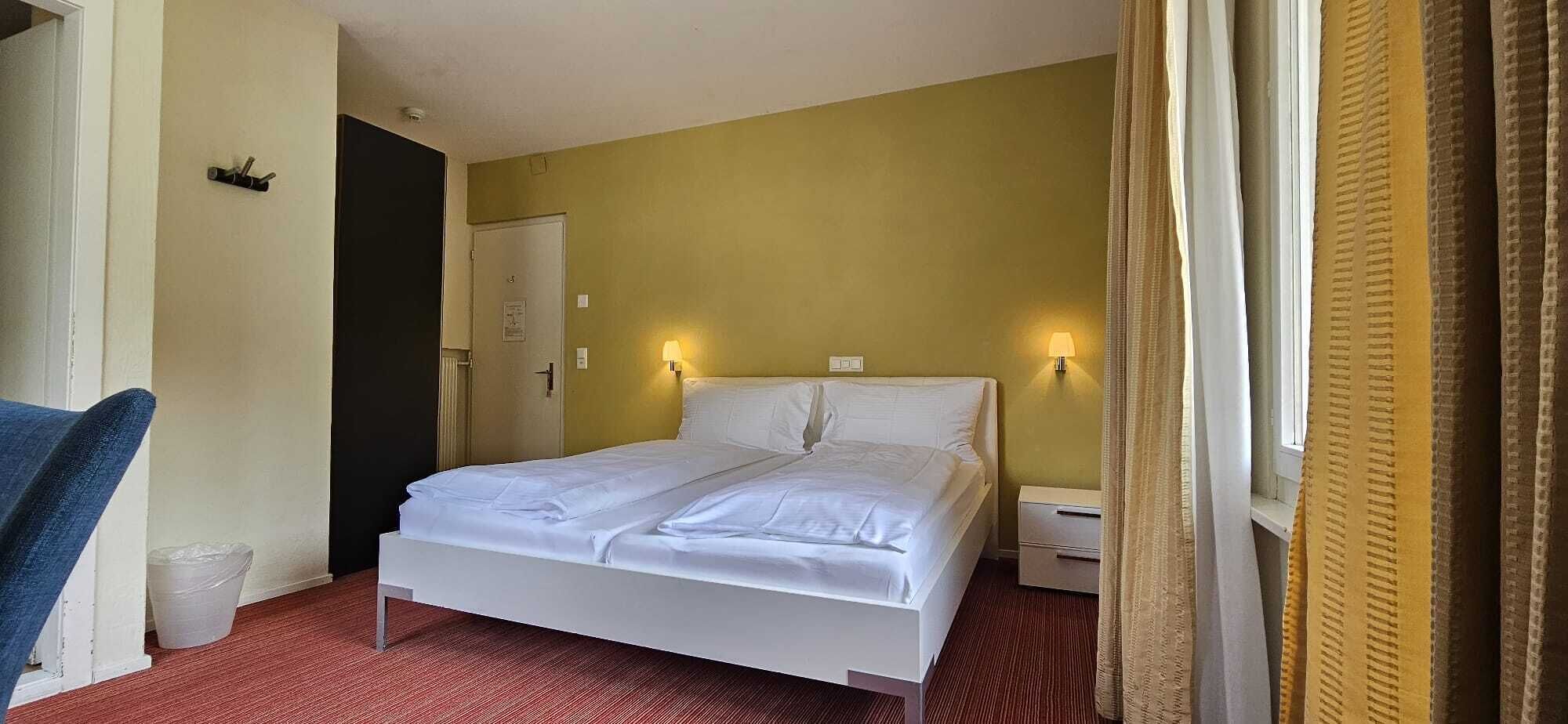 A hotel room with a bed, a chair, a nightstand, and a door.