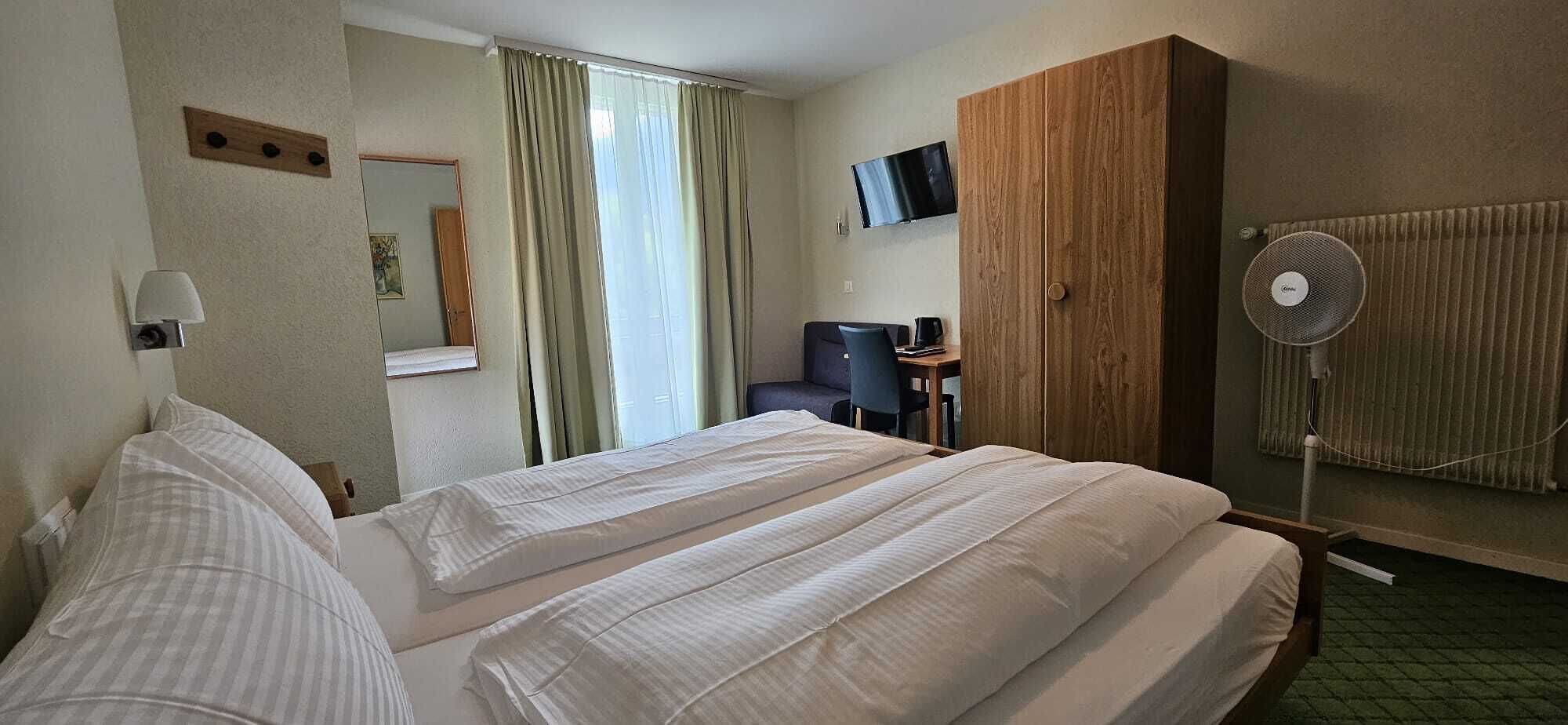 A hotel room with a large bed, a desk with a chair, a TV mounted on the wall, and a standing fan.