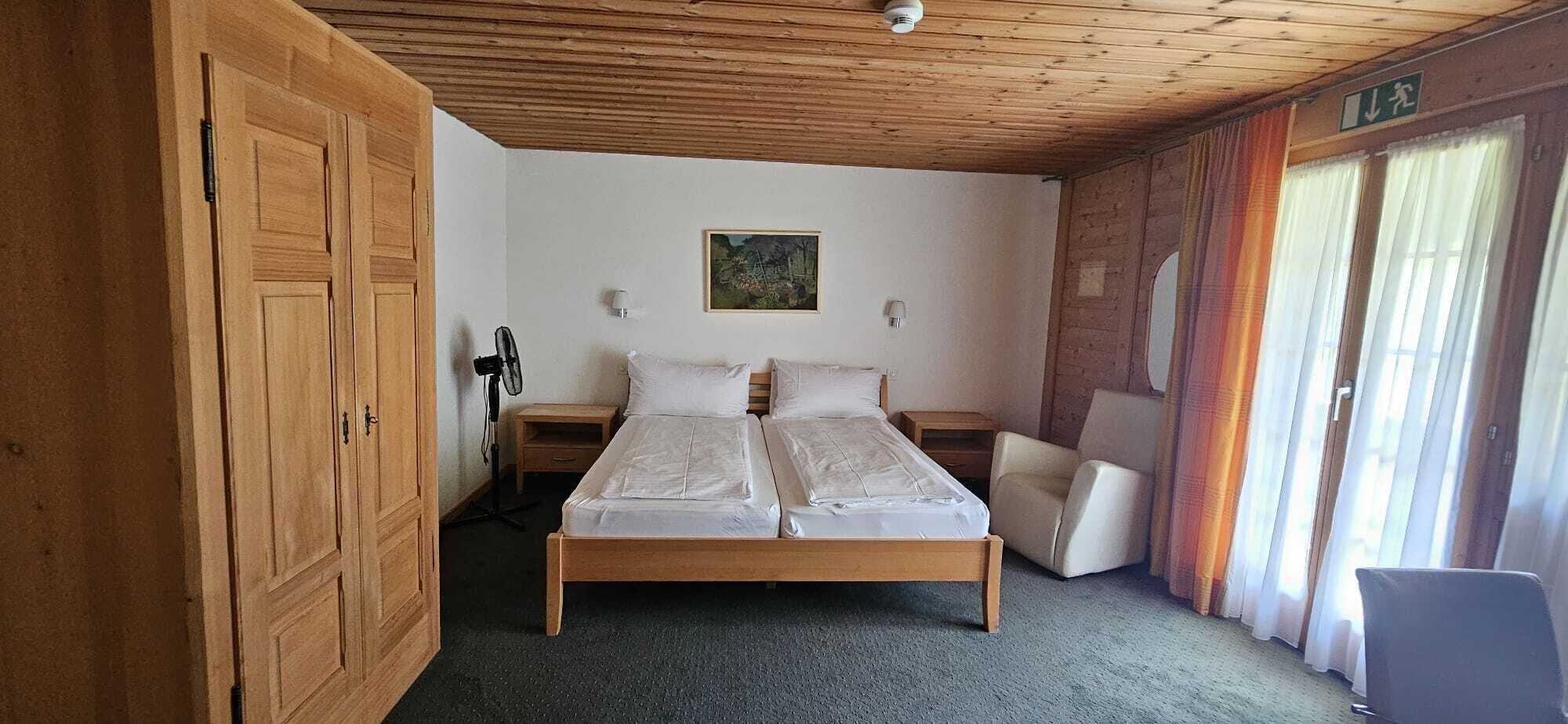 Une chambre avec un lit en bois, un ventilateur, une chaise et un tableau accroché au mur.