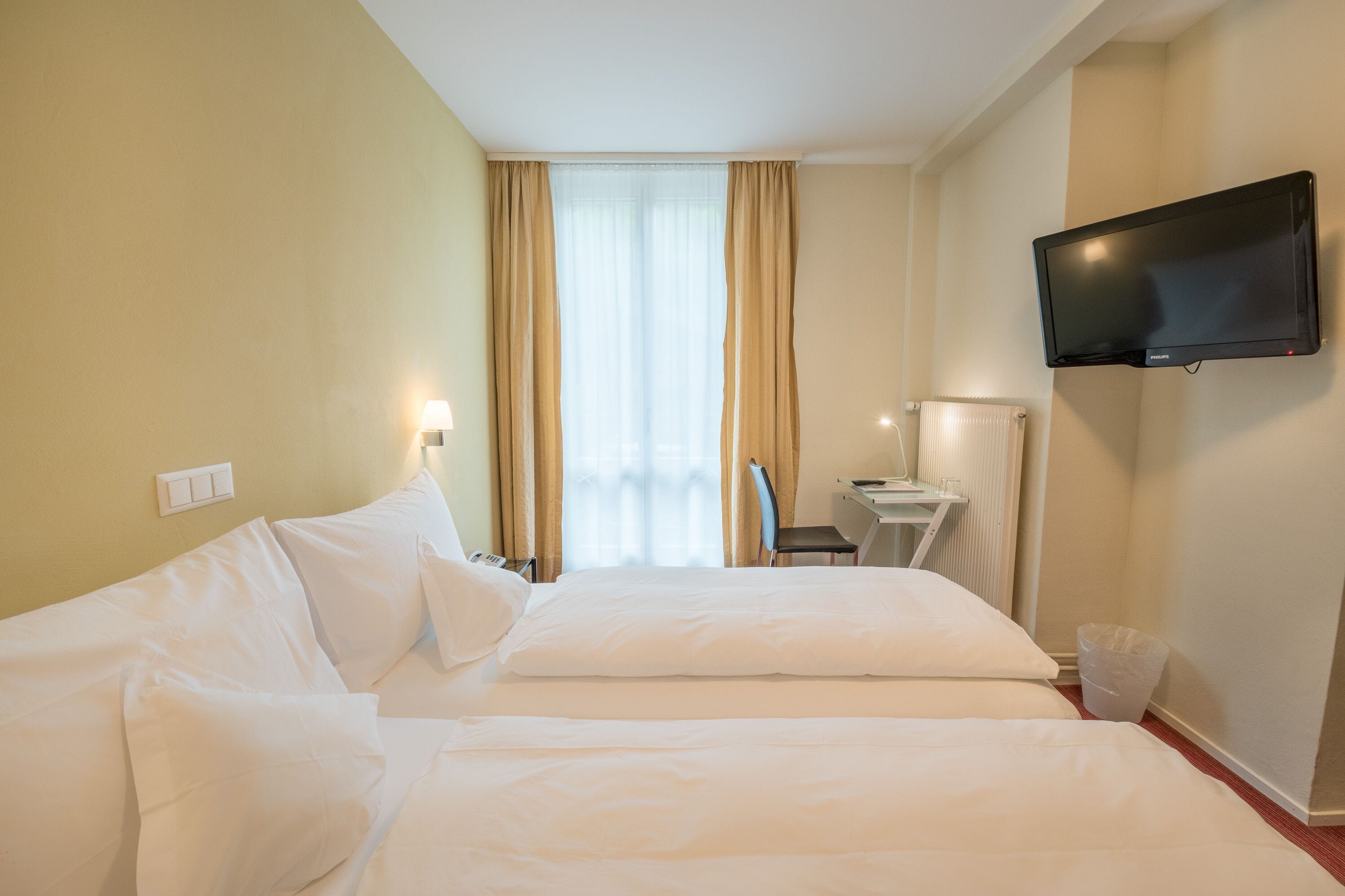 Habitación de hotel con dos camas, un televisor montado en la pared, un escritorio con una silla y una ventana con cortinas.