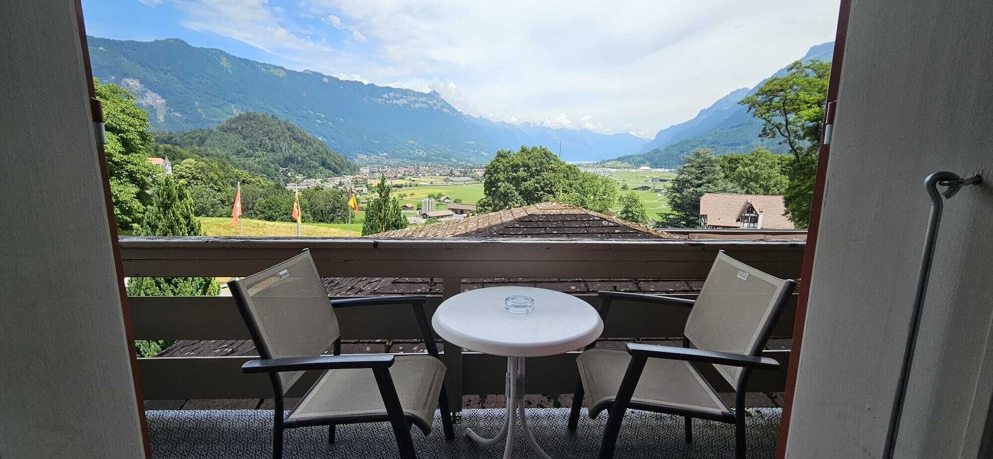 A balcony with two chairs and a small table, overlooking a mountainous landscape with a village below.