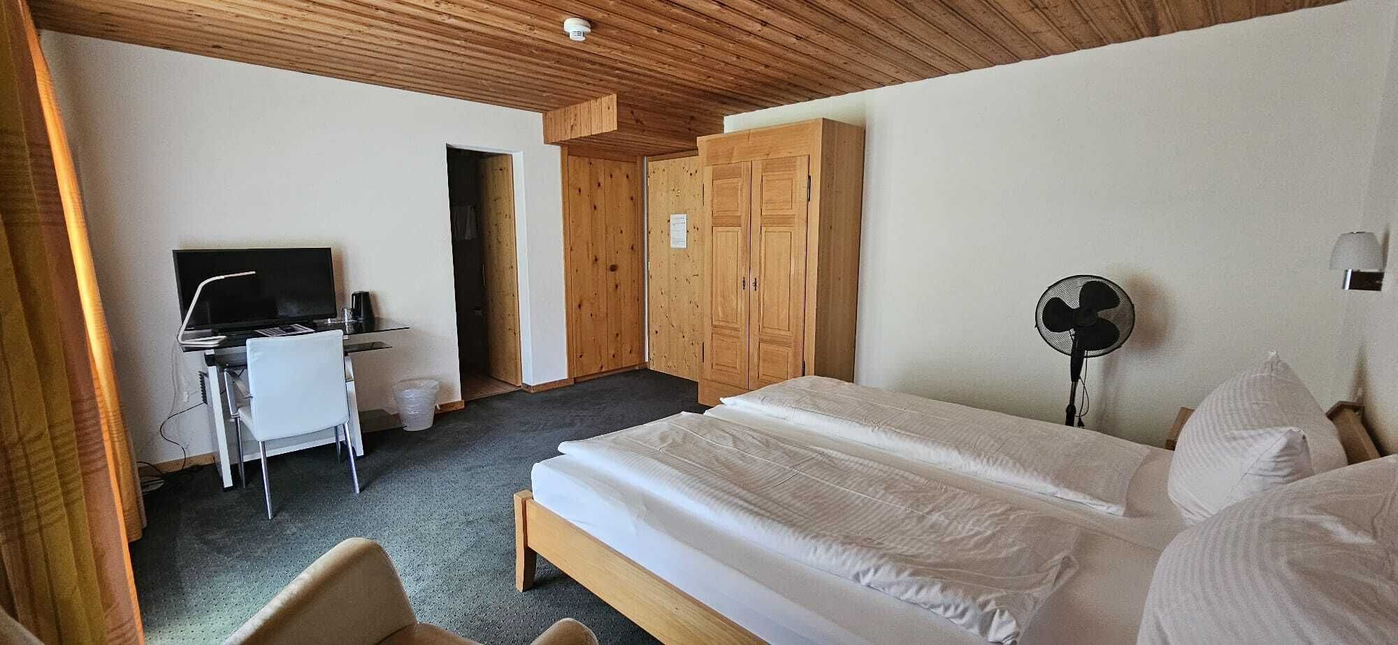 A hotel room with a bed, a desk with a TV, a fan, and wooden wardrobes.