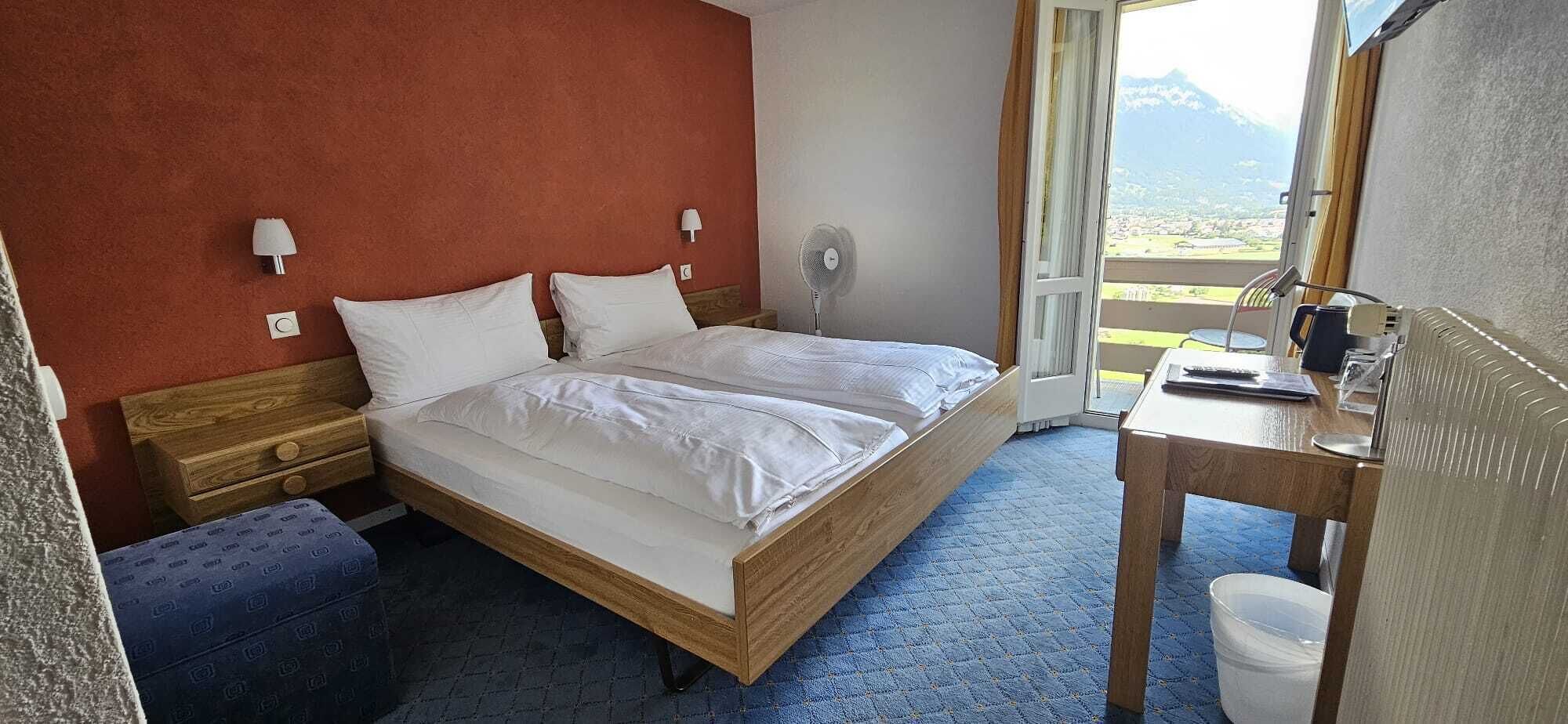 Ein Schlafzimmer mit einem hölzernen Bett, Nachttischen, einem Schreibtisch und Blick auf Berge durch das Fenster.