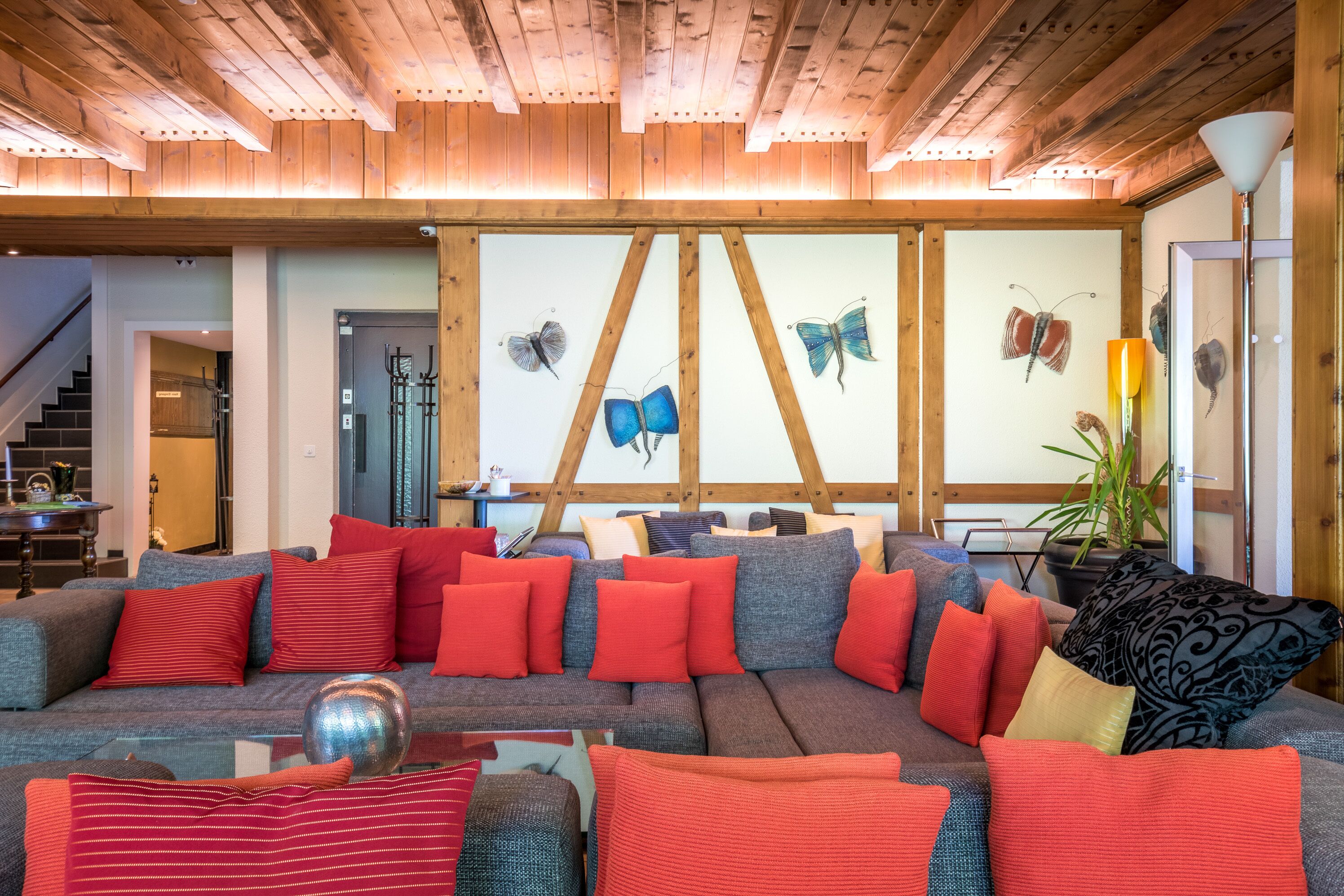 A cozy living room with a large sectional sofa, red and grey cushions, wooden ceiling beams, and butterfly wall art.