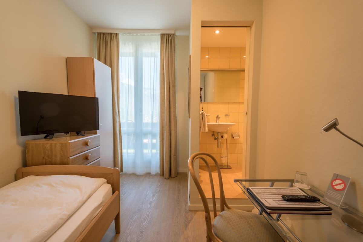 Habitació d'hotel amb llit, televisió, escriptori amb cadira, bany amb mànec i mirall, i finestra amb cortines.