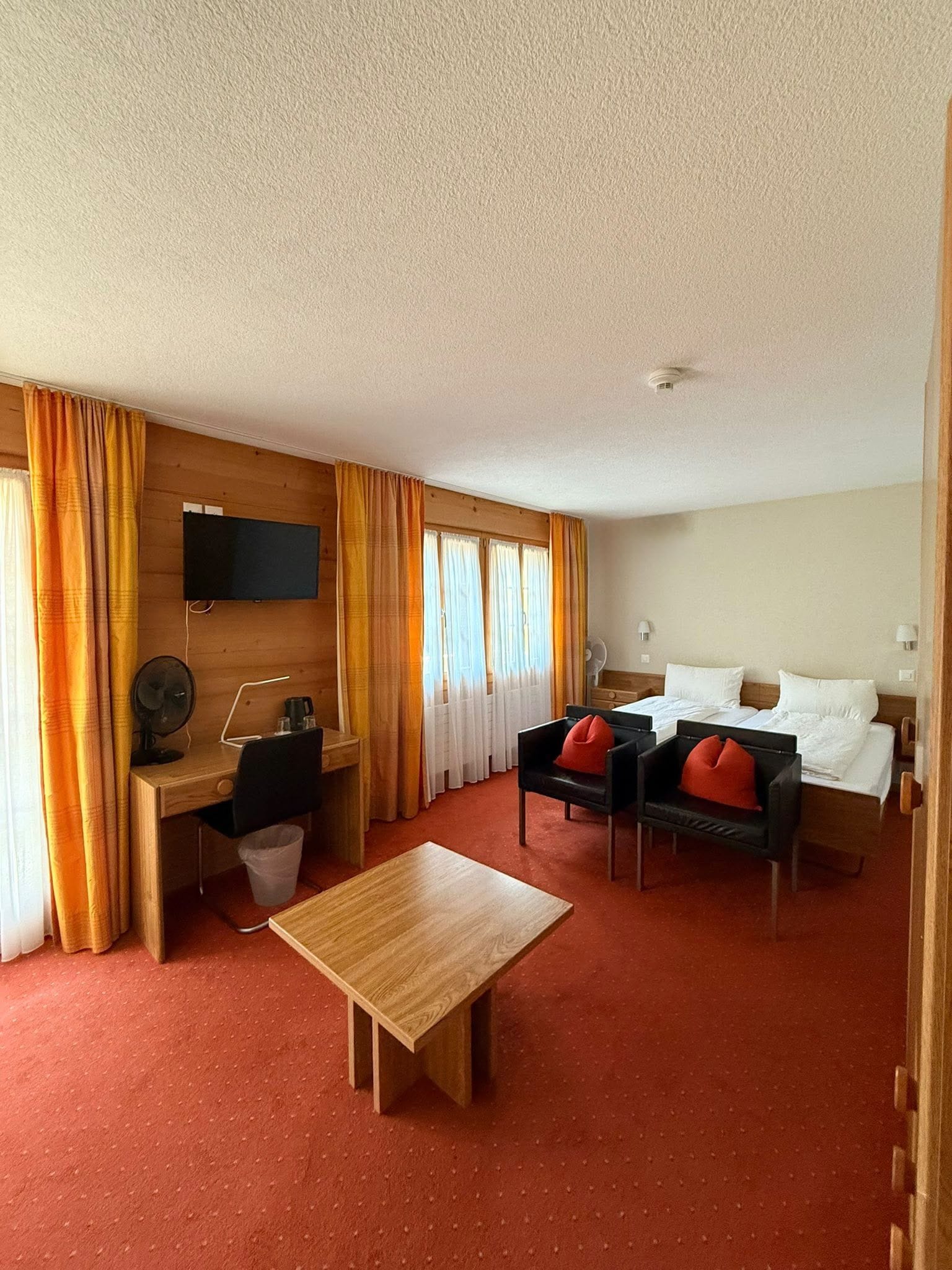A hotel room with a bed, a desk, a chair, a small table, and a TV mounted on the wall.