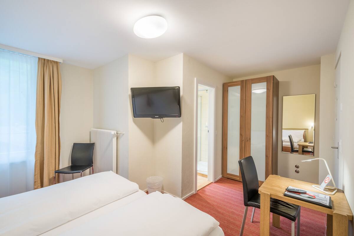 Habitació d'hotel amb llit, escriptori amb cadira, televisió, mirall i finestra amb cortines.