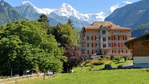 Un hôtel de style architectural européen traditionnel, entouré d’une végétation luxuriante et avec des montagnes en toile de fond.