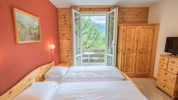 A bedroom with a wooden bed, a painting on the wall, a window with a view of greenery and mountains, and a wooden wardrobe.