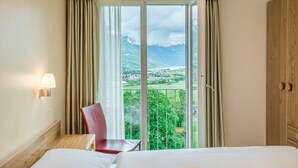 Une chambre d’hôtel avec un lit, une chaise, une table, une lampe et une grande fenêtre avec des rideaux.