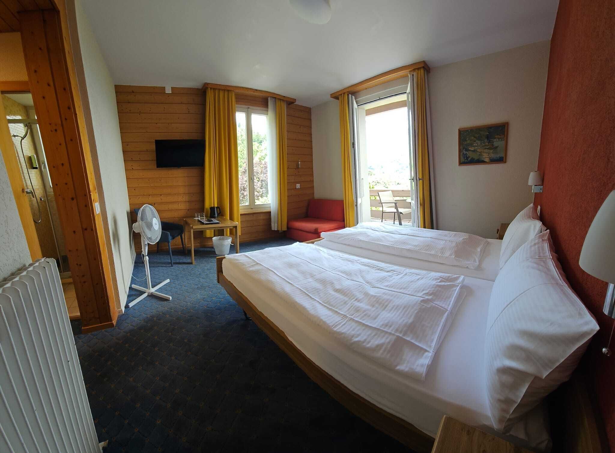 A hotel room with a double bed, a small table with a chair, a fan, and a window with curtains.