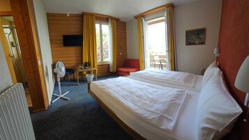 A hotel room with a double bed, a small table with a chair, a fan, and a window with curtains.
