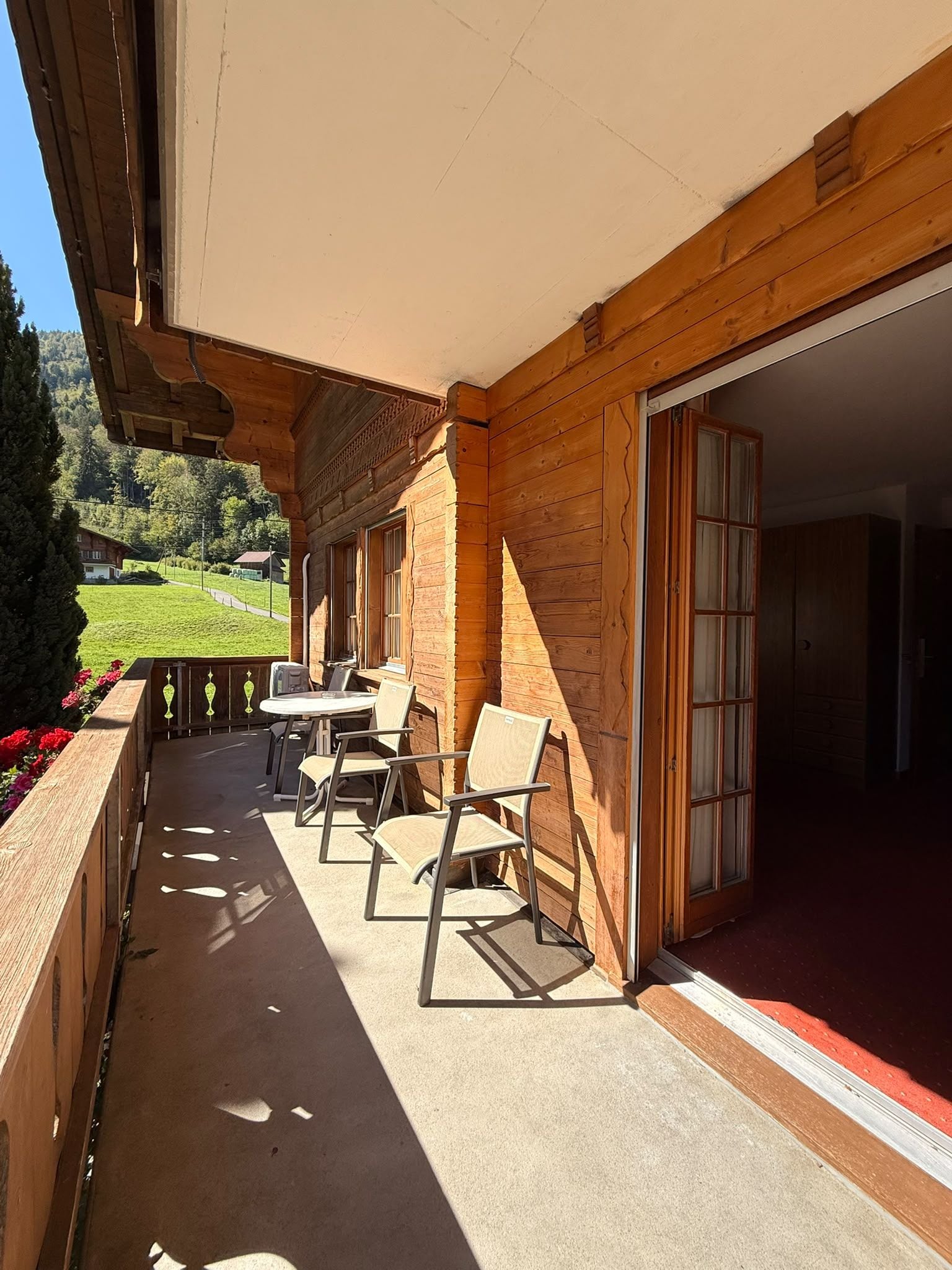 A wooden building with a covered outdoor area, a table with chairs, and a view of a green landscape.