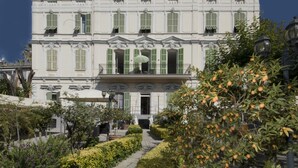 Un edificio grande de varios pisos con una fachada simétrica, una entrada central y un jardín cuidado con diversas plantas y un camino que lleva a la entrada.