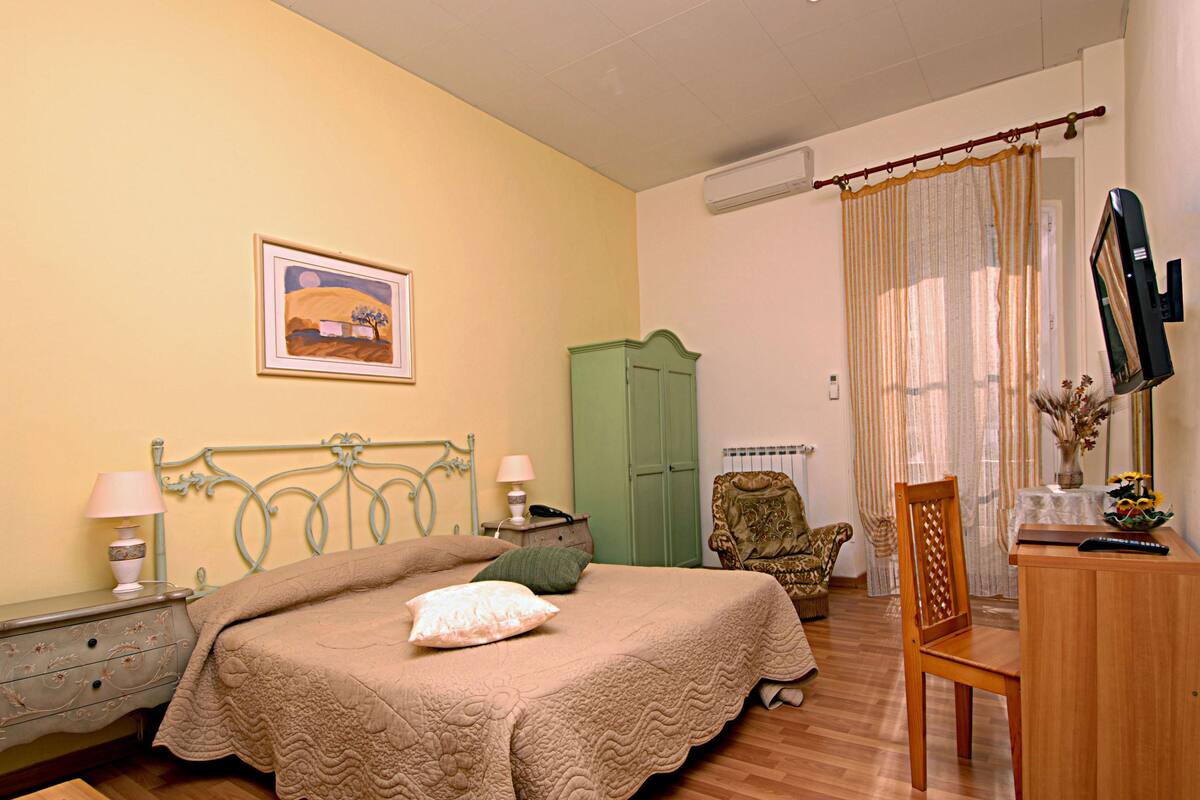 A bedroom with a bed, a green wardrobe, a wooden chair, a desk with a vase, and a television.