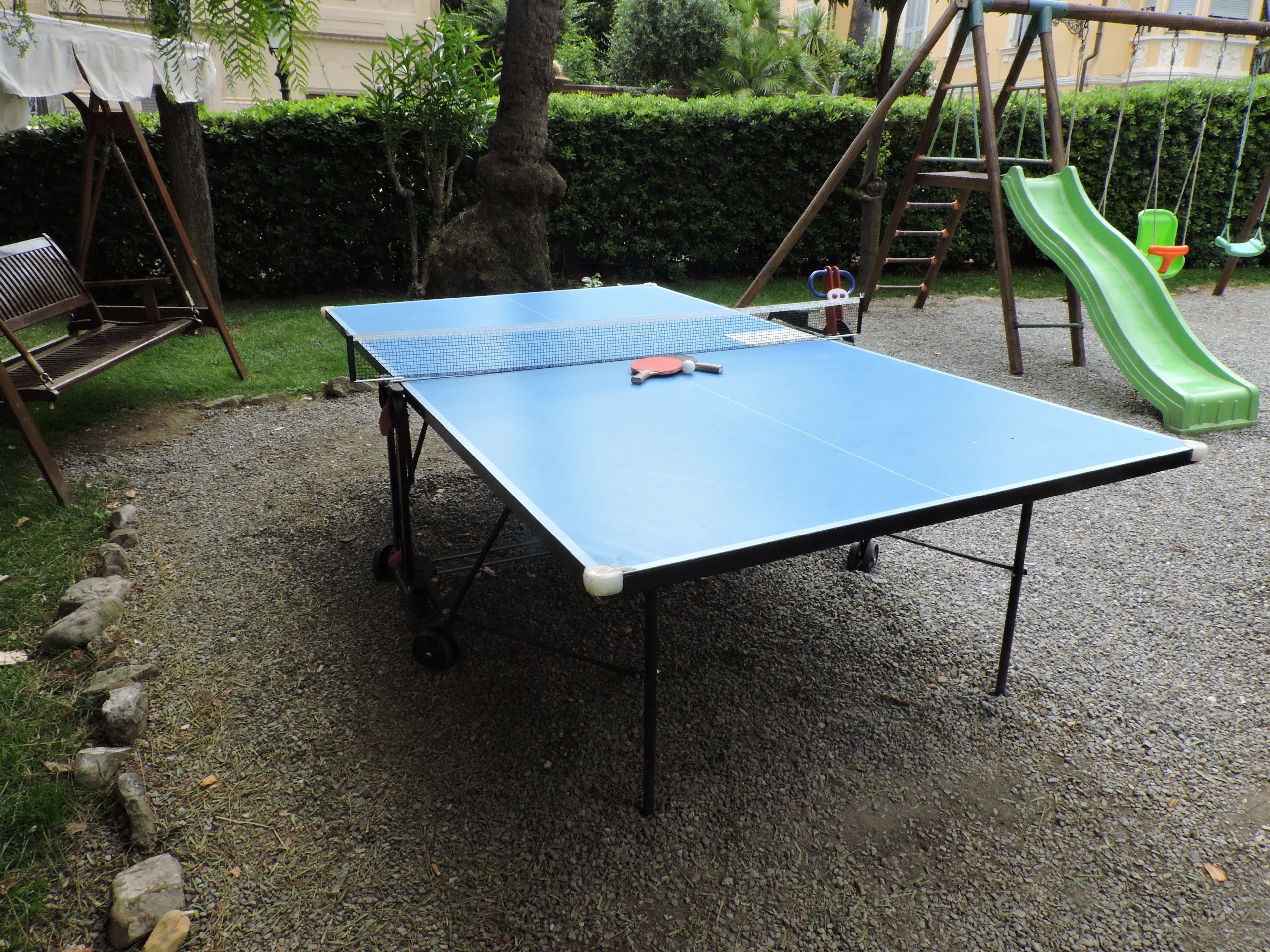 Una mesa de ping pong en un espacio exterior con un parque infantil al fondo.