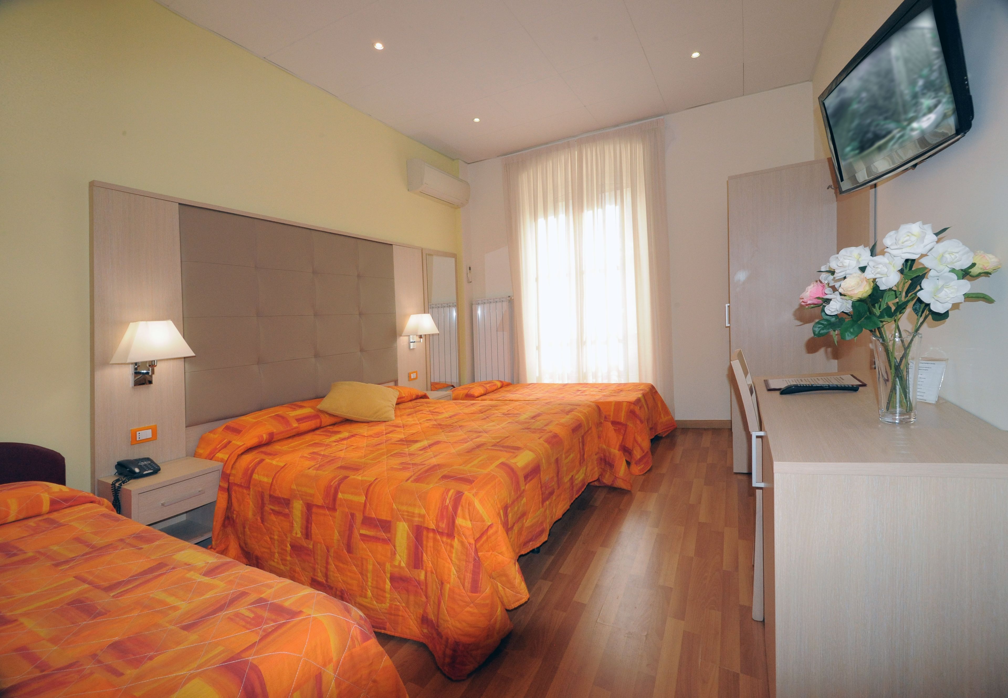 A hotel room with two beds, a desk, a TV, and a vase with flowers.