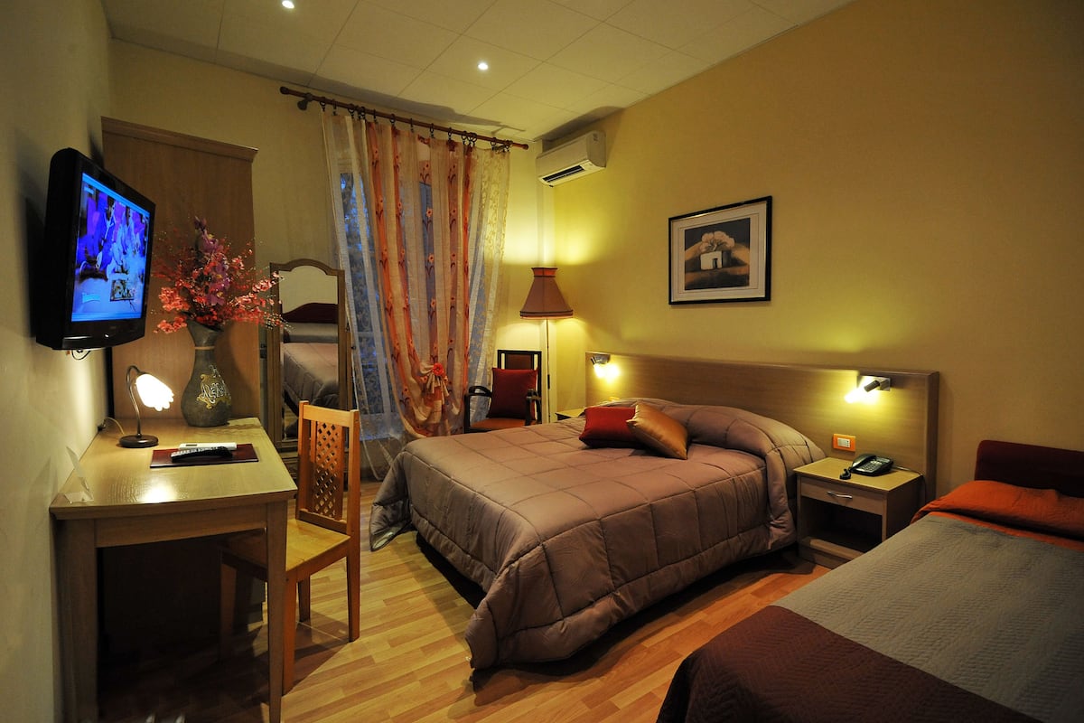 A hotel room with a bed, a desk with a lamp, a TV, and a chair.