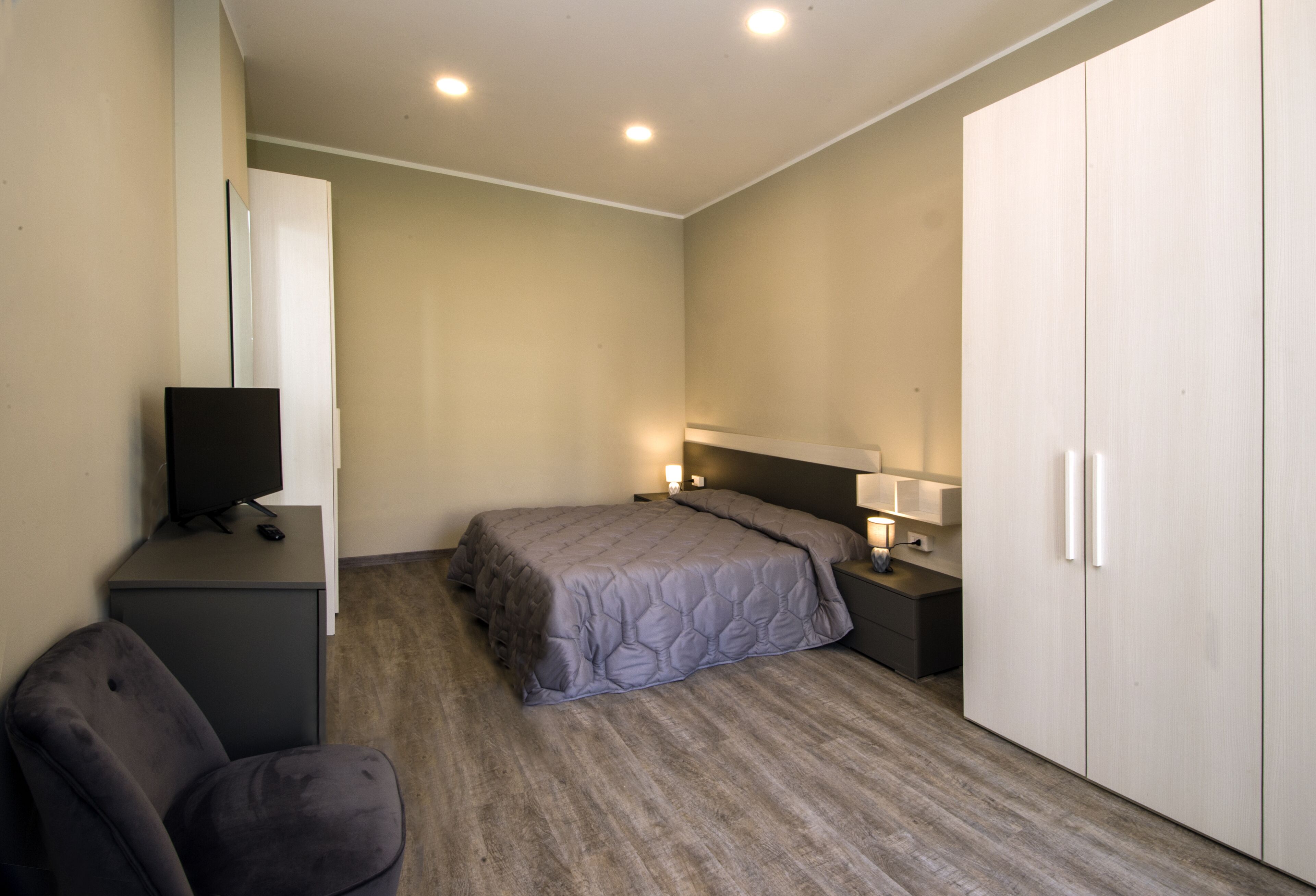 A bedroom with a bed, a desk with a computer, a chair, a wardrobe, and bedside lamps.
