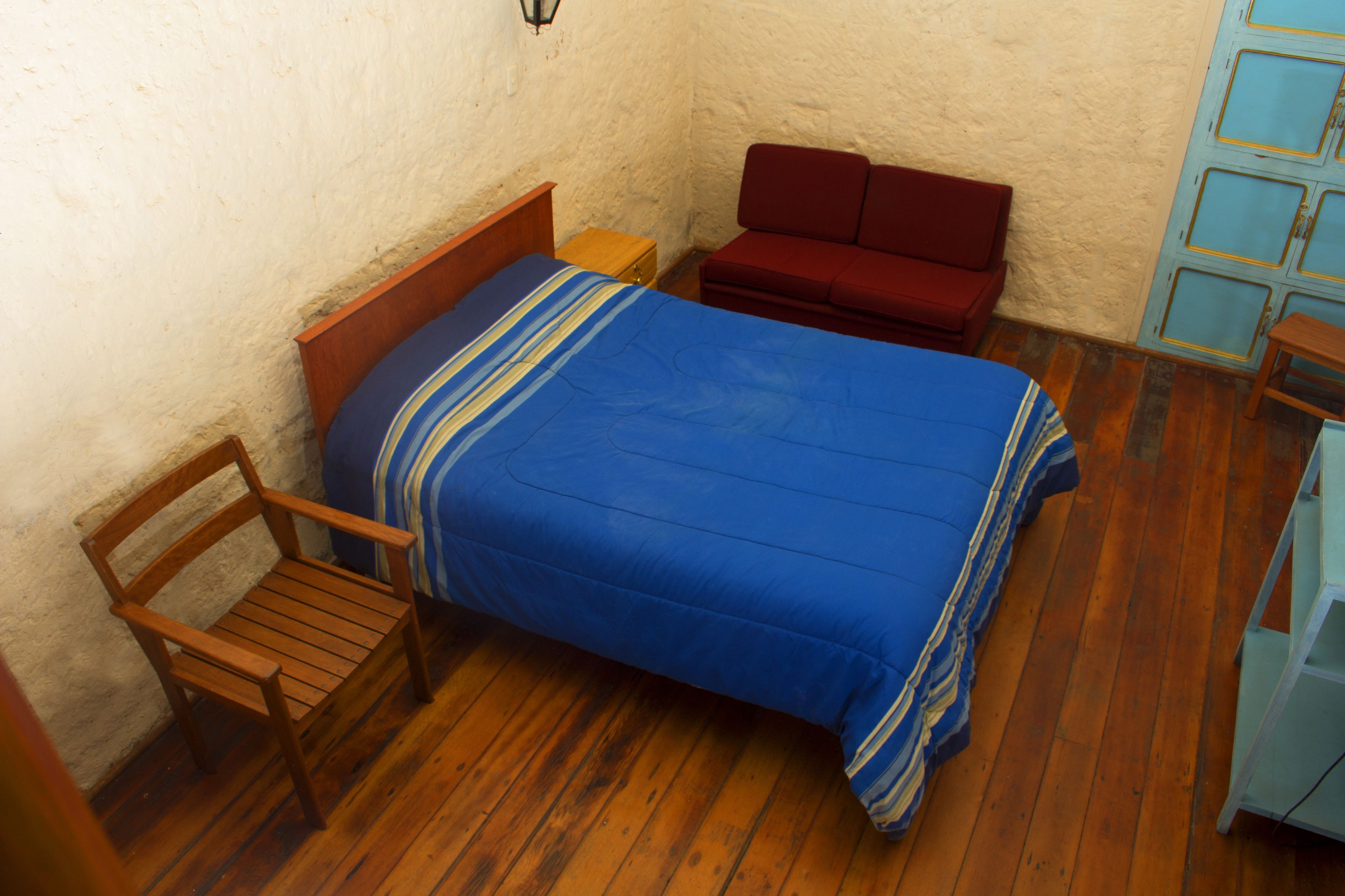 Ein kleiner, gemütlicher Raum mit Holzboden, einem Einzelbett mit blauer Bettdecke, einem Holzstuhl, einem roten Sofa und einem kleinen Holztisch.