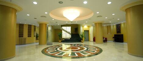 A spacious lobby with a central circular floor design, multiple columns, and a staircase leading to upper levels.
