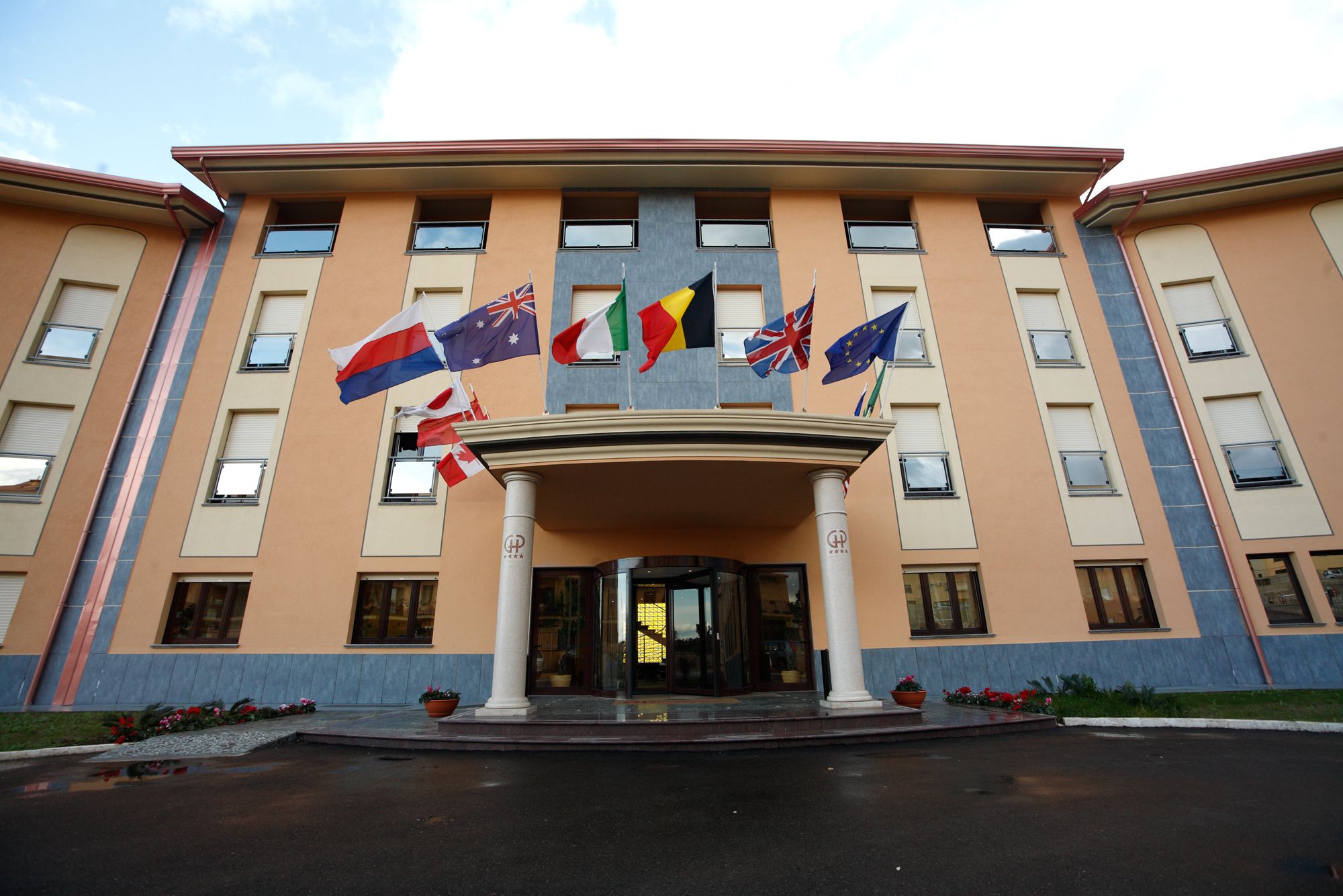 A multi-story building with a central entrance, multiple windows, and a variety of flags displayed.