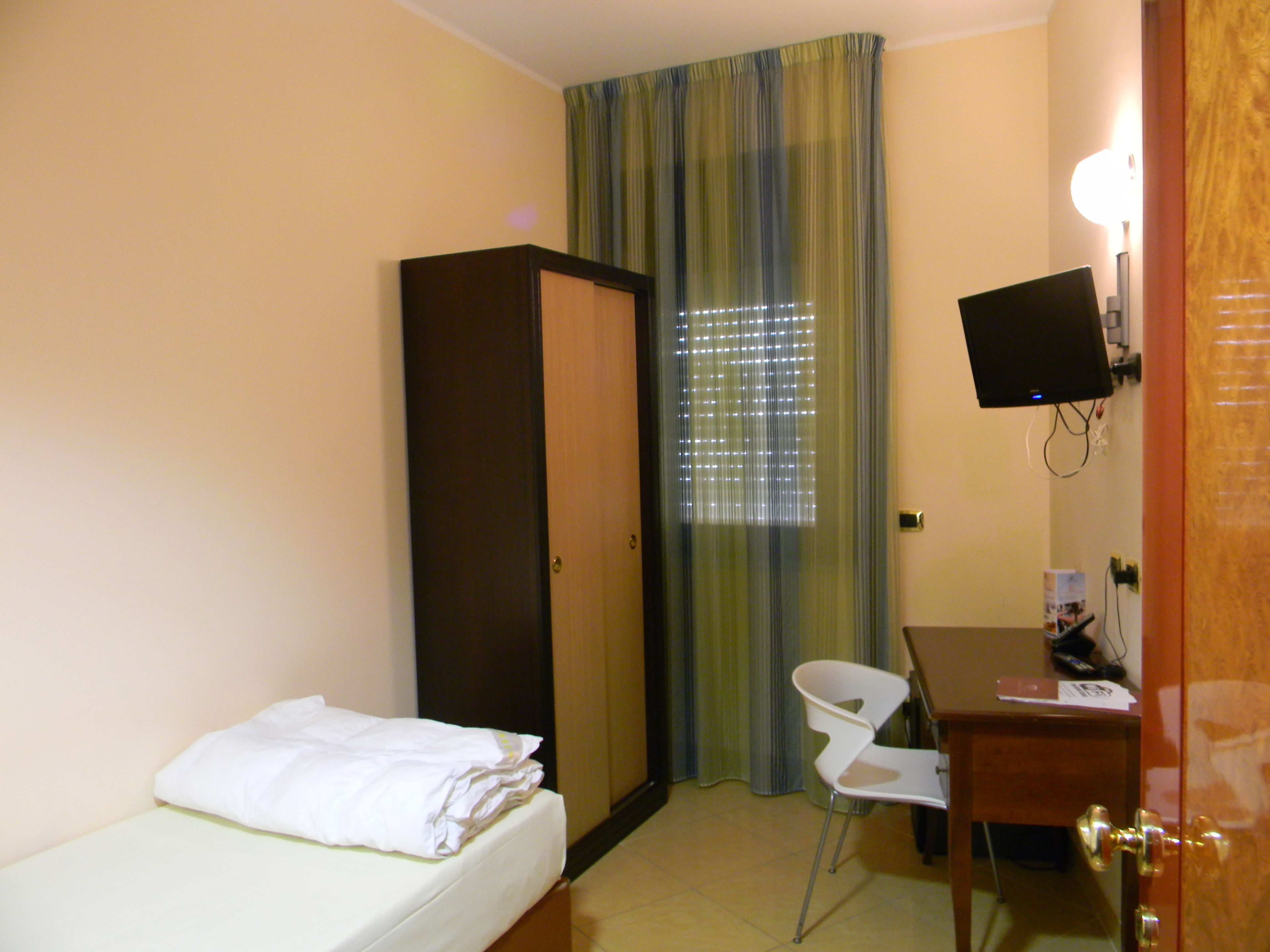 Habitación de hotel con cama, escritorio, silla, armario, televisor y ventana con persianas.