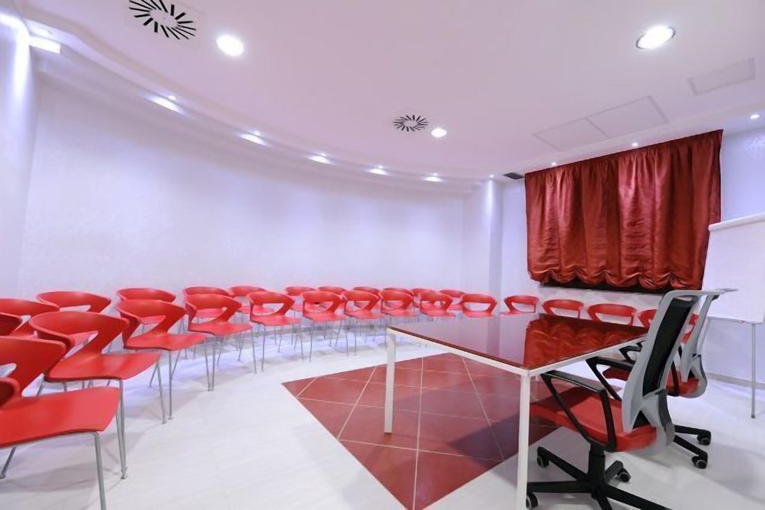 Ein Konferenzraum mit einem langen Tisch und roten Stühlen, weißen Wänden und einem roten Vorhang.