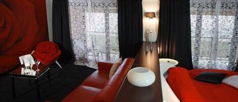 A modern hotel room with a red sofa, a wooden side table, and a large window with black curtains.