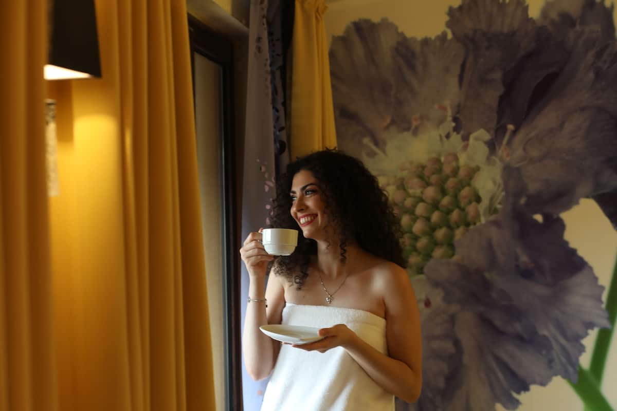 A person in a white towel holding a cup and a plate, standing in front of a window with yellow curtains and a large floral painting.