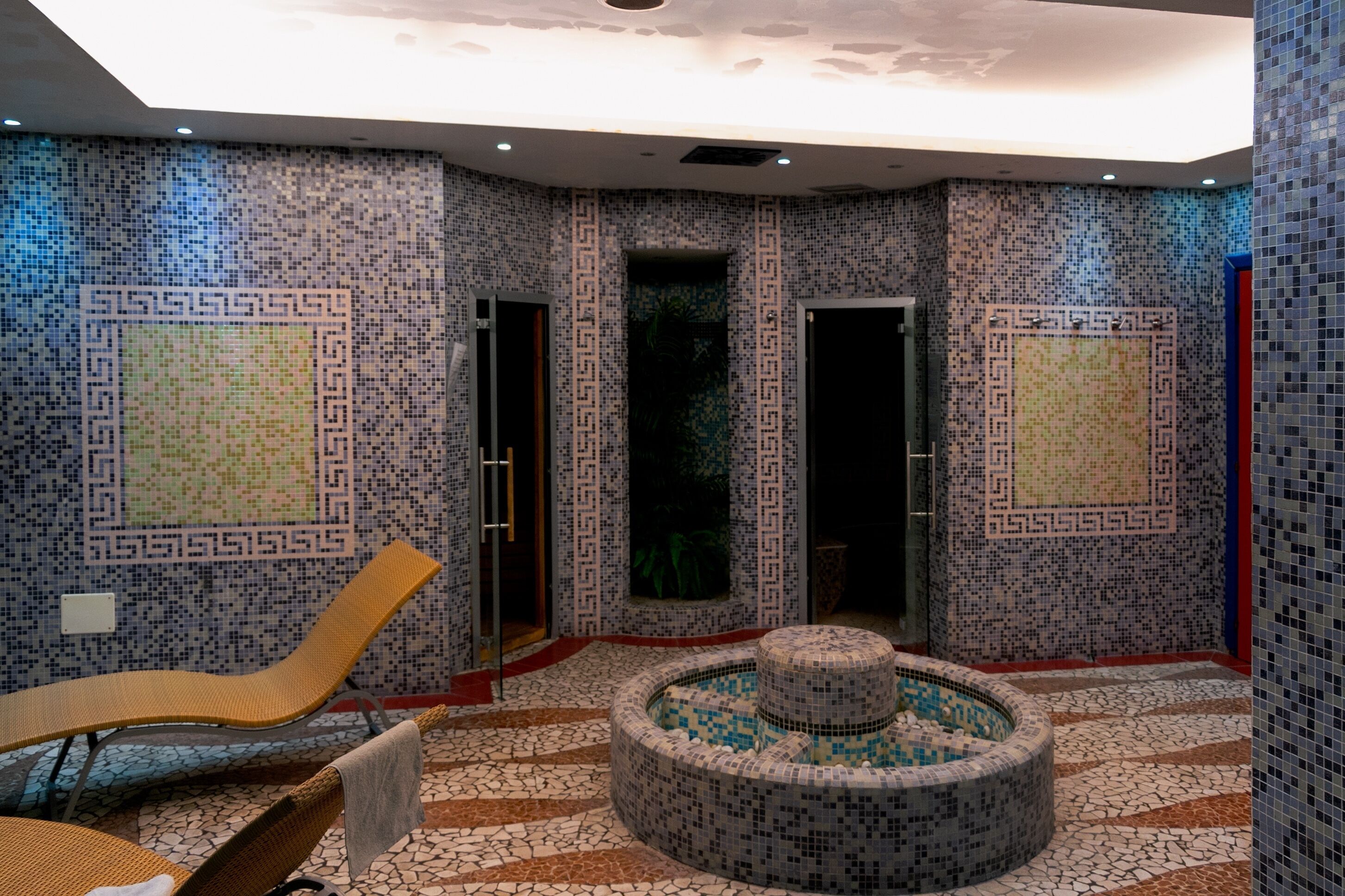 Uma área de spa com paredes em mosaico, um elemento de água circular e uma cadeira de lounge.