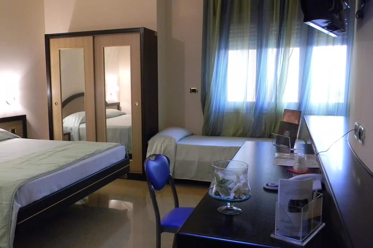 A hotel room with a bed, a desk with a chair, a small table, a glass, and a wardrobe.