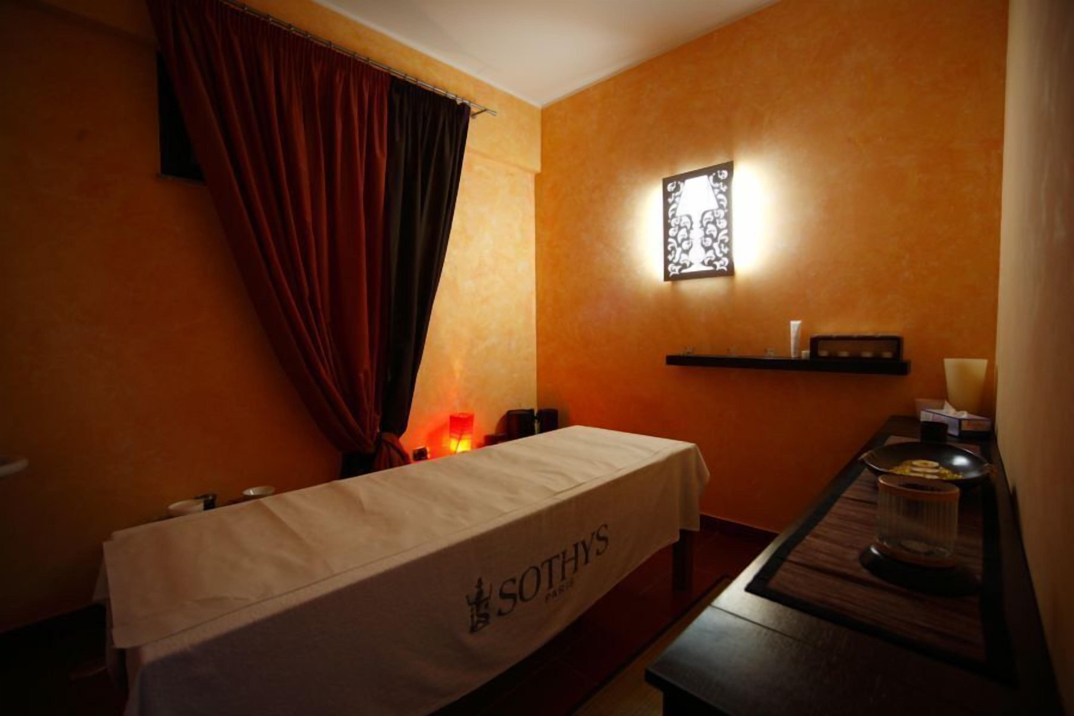 A room with a table covered by a cloth, a wall-mounted shelf with a candle, and a window with curtains.