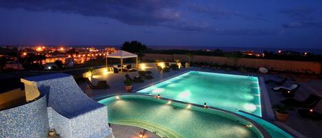 A swimming pool with a tiled deck, lounge chairs, and a view of the city at dusk.